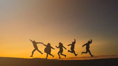 Five silhouetted figures jumping against a vibrant sunset sky with visible horizon.