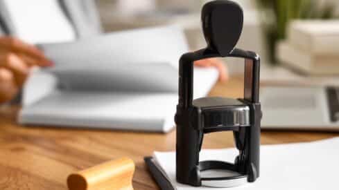 Black self-inking stamp on desk with open notebook and hand, suggesting office setting.