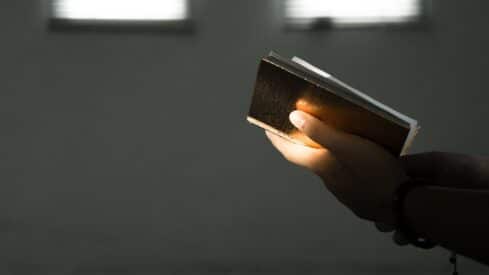 Hand holding an open book with a dark cover, softly lit in a dim environment.