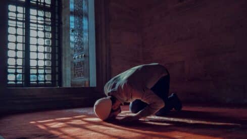 Person kneeling in dim room, praying on carpet; sunlight through lattice window creates shadows.