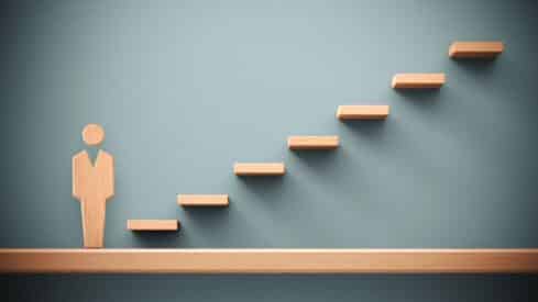 Wooden figure next to ascending wooden steps on a greenish-blue wall, symbolizing growth.