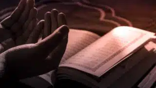 Hands raised in prayer beside an open book with Arabic script in soft light.