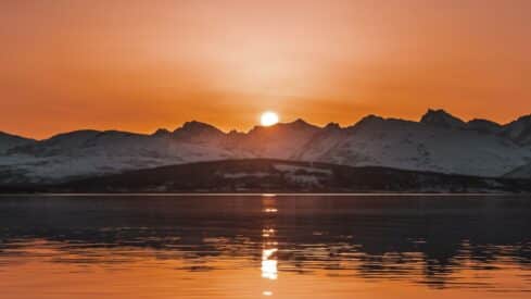 Sunset over snowy mountains, reflecting on a calm lake, in warm orange colors.