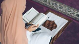 Person in peach headscarf reading a Quran with Arabic script on a patterned prayer mat.