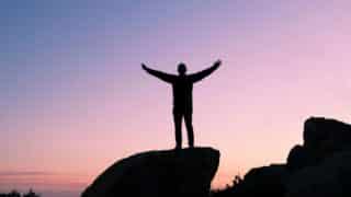 Silhouetted figure with outstretched arms on rock against pastel sky at dusk or dawn.