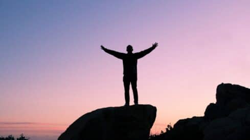 Silhouetted figure with outstretched arms on rock against pastel sky at dusk or dawn.