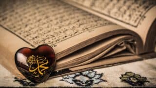 Ornate holy Quran with Arabic script open on fabric, heart-shaped object with calligraphy on top.