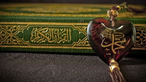 Heart-shaped pendant with Arabic calligraphy in front of a green holy Quran with Arabic script.