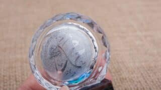 Decorative glass object with Arabic calligraphy emblem held in fingers against neutral background.