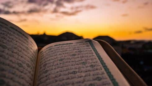 Open Quran with Arabic text, sunset sky and silhouetted buildings in the background.