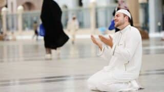 Person in white garment and cap kneeling in prayer in open public space with blurred figures.