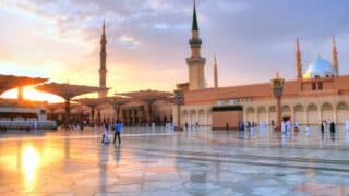 Mosque courtyard at sunset with minarets, domes, umbrellas, and people on marble pavement.