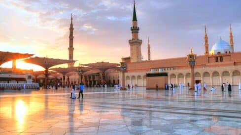 Mosque courtyard at sunset with minarets, domes, umbrellas, and people on marble pavement.