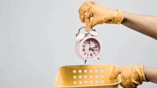 Hands in yellow gloves hold a pink alarm clock above a perforated yellow basket.