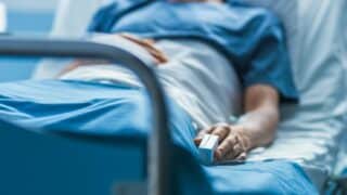 Person in hospital bed holding a pulse oximeter on their hand, wearing a blue gown.