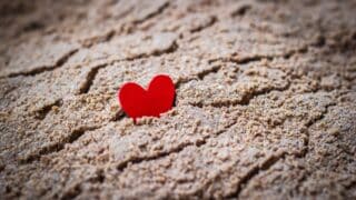 Small red heart partly embedded in cracked, textured brown sand surface.