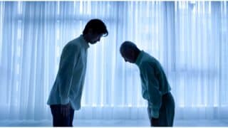 Two people bow to each other in a room with sheer curtains and bluish lighting.