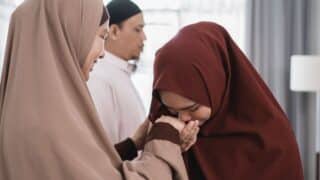 Two women in hijabs interacting; one kisses the other's hand, man in background, indoor setting.