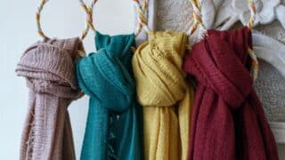 Four scarves in brown, teal, yellow, and red on decorative hangers against a light backdrop.