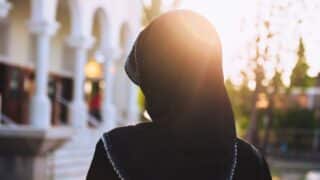 Person in black headscarf with sunlight lens flare, arched structures in soft-focus background.