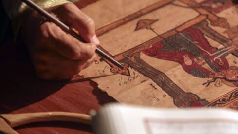 Person detailing art with brush on parchment with intricate patterns and traditional figure.