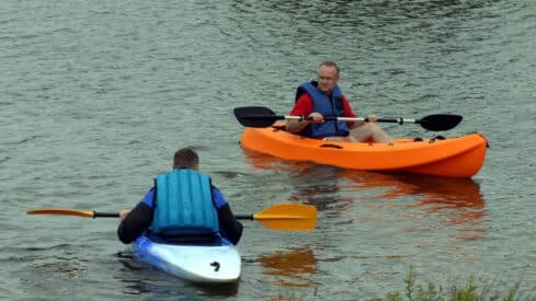 Two people kayaking, wearing blue life jackets. Orange, blue, and white kayaks. Holding paddles.