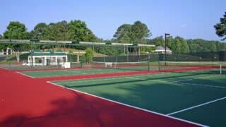 Outdoor tennis court complex, red-green surfaces, trees, clear sky, small building nearby.