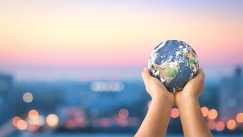 Hands holding a small globe against a blurred cityscape at sunset with pink and blue hues.