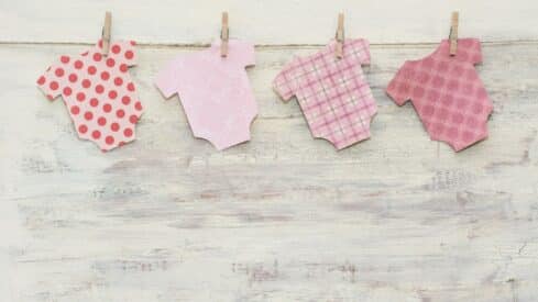 Paper cutouts of pink baby onesies clipped to a line against a rustic wooden background.