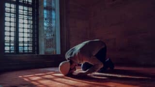 Person praying on a carpet in a dimly-lit room near a window with intricate ironwork.