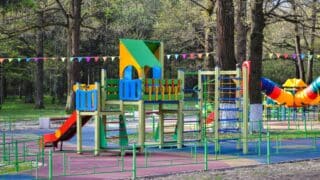 Colorful playground with slides, climbing frames, tunnels, trees, bunting, and green fencing.