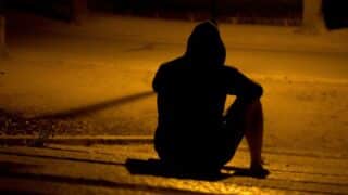 Person in hoodie sitting on steps at night, silhouetted by warm streetlight.