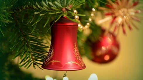 Red bell ornament with gold patterns on Christmas tree branch, blurred decorations behind.