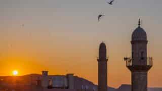 Sunset over hills with two minarets, birds in sky, and rooftops at bottom.