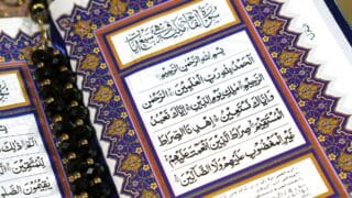 Open Quran page with ornate calligraphy, blue and gold borders, black prayer beads.