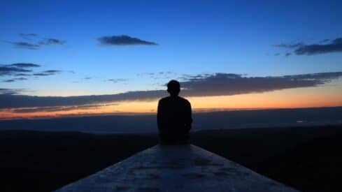 Person sitting on ledge facing a vast sunrise or sunset landscape, with colorful sky and clouds.