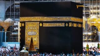 Kaaba draped in black and gold, surrounded by a crowd and scaffolding indicating construction.