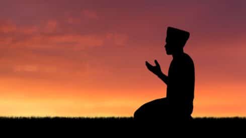 Silhouette of a kneeling person with hands raised in prayer against a vibrant sunset.