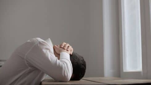 Person in white shirt resting head on table, hands clasped, indoor near large window.