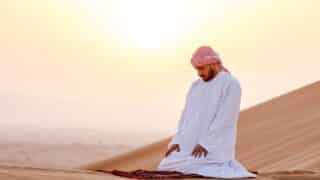 Person kneeling on rug in desert at sunset, wearing traditional attire, sand dunes in background.