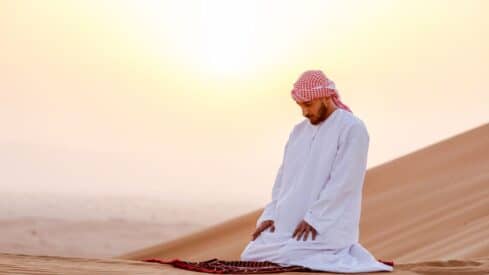 Person kneeling on rug in desert at sunset, wearing traditional attire, sand dunes in background.