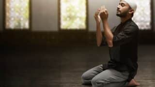 Person kneeling in dim room, wearing black outfit, cap, praying with raised hands.