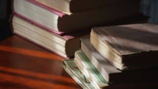 Stack of old hardcover books with worn pages on a wooden surface, lit by natural light.