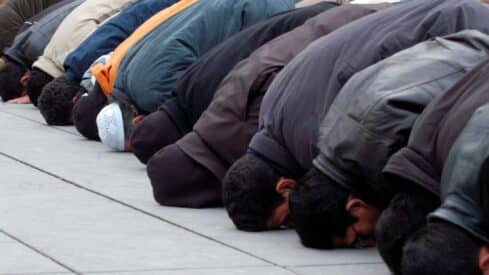 People kneeling in prayer outdoors, foreheads touching ground, wearing jackets and coats.