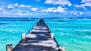 Wooden pier over turquoise water leading out to sea under a partly cloudy blue sky.