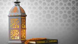 Ornate lantern with warm glow beside book and prayer beads, geometric pattern backdrop.