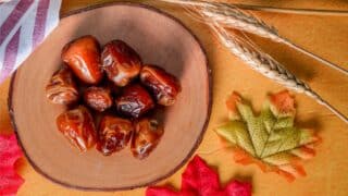 Wooden bowl with dates on orange surface, surrounded by wheat stalks and autumn leaves.