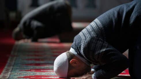 Individuals praying on a mosque carpet, kneeling with foreheads down, wearing dark clothing.