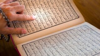 Person points at holy Quran in Arabic text in book with prayer beads, on a wooden surface.