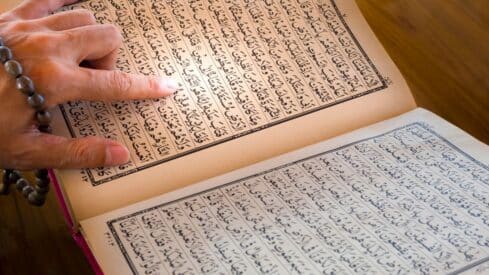 Person points at holy Quran in Arabic text in book with prayer beads, on a wooden surface.